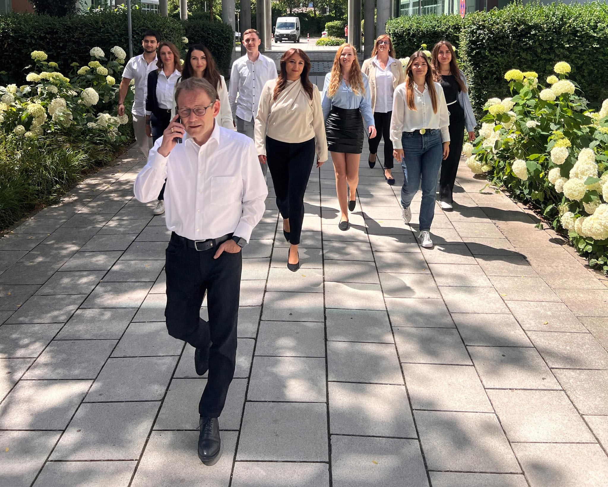 Ein Mann telefoniert voran, gefolgt vom Team der Lkm GmbH auf einem Gartenweg mit weißen Blumen. Steuerberatung Unterföhring.