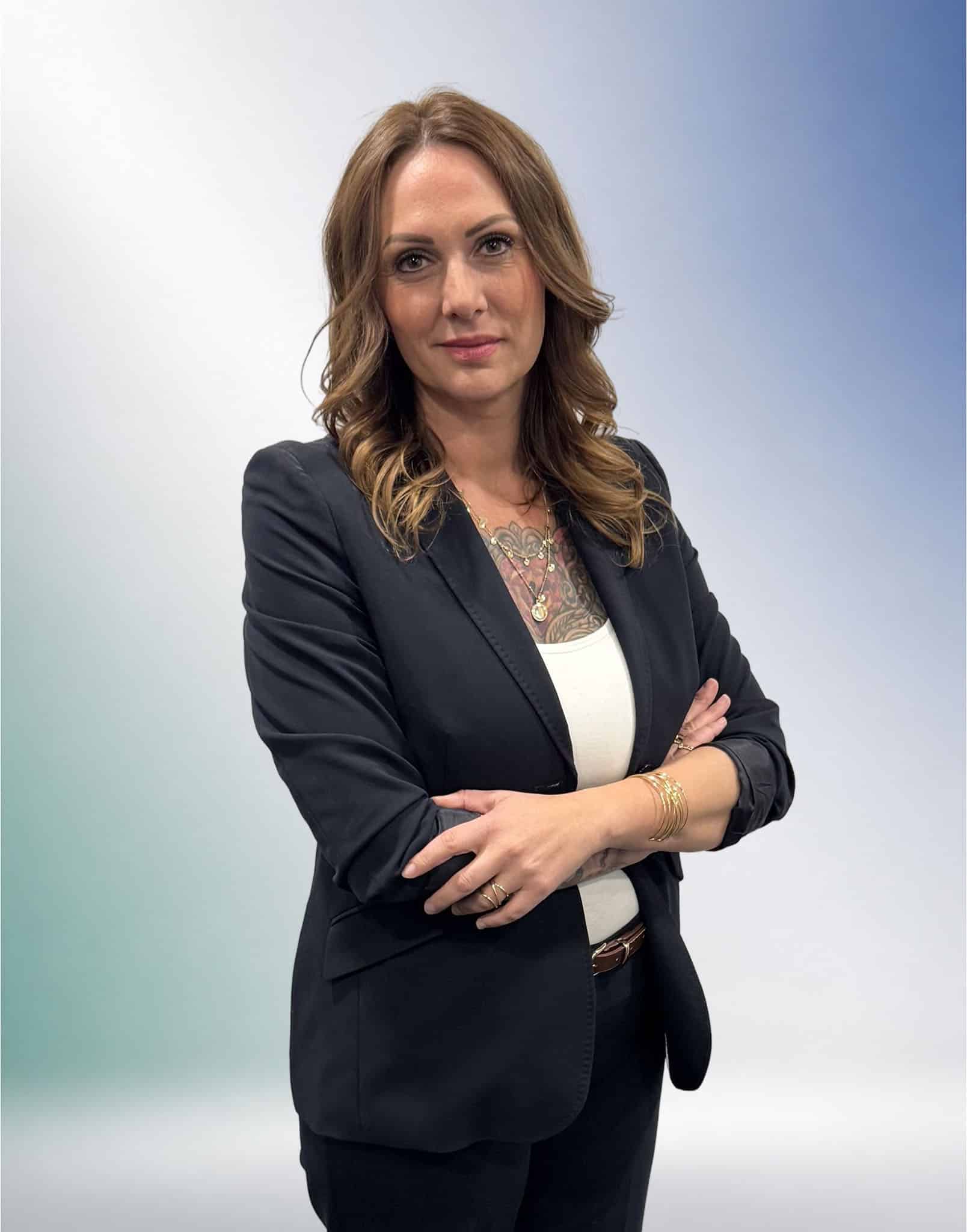 Selbstbewusste Frau mit Tattoos in einem schwarzen Blazer vor einem grün-blauen Farbverlauf, Rechtsanwältin für Rechtsberatung in Unterföhring.