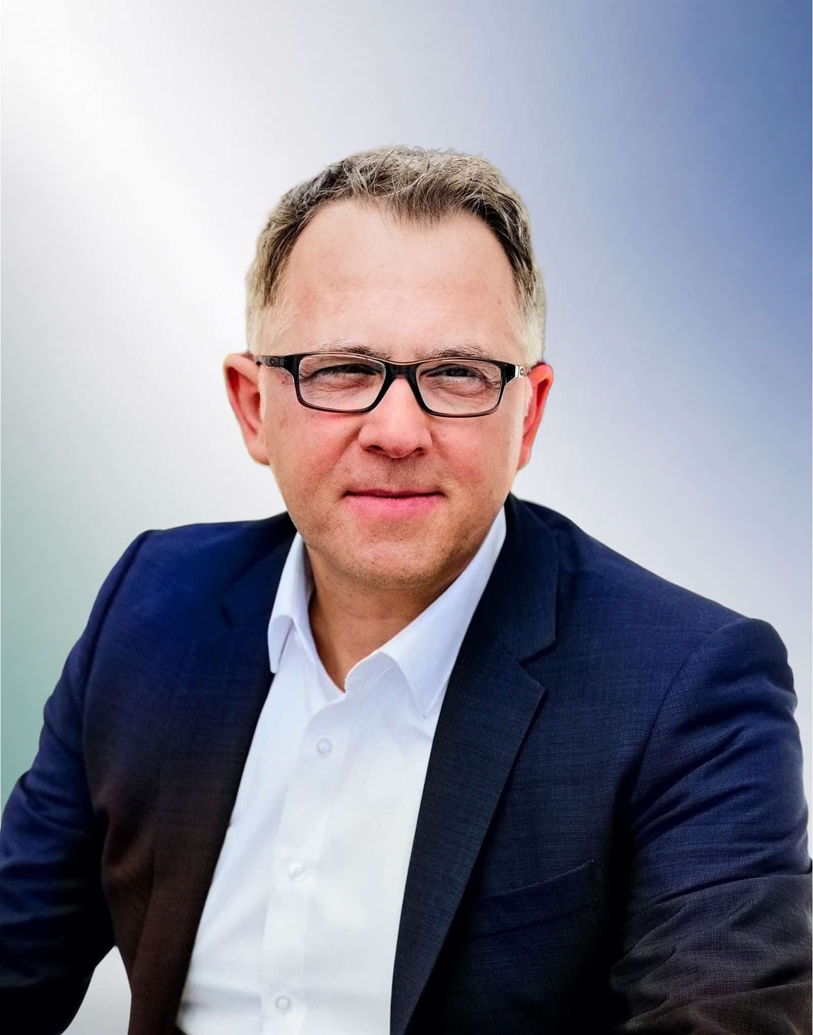 Mann mit Brille in einem dunklen Sakko vor einem Farbverlauf, mit ernstem Blick – Anwaltskanzlei München, Rechtsanwalt in Unterföhring.