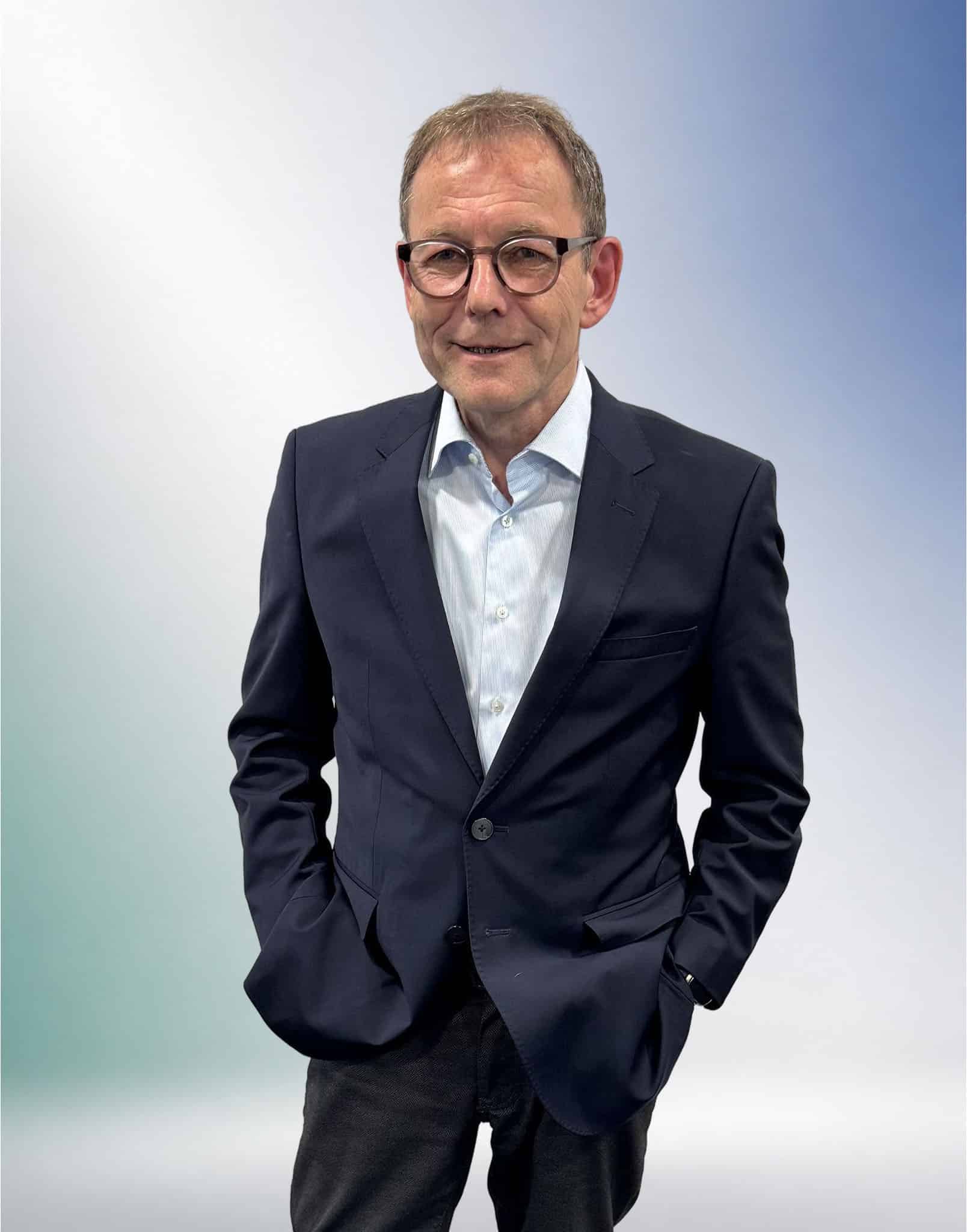 Ein Mann mit Brille im blauen Anzug steht mit den Händen in den Taschen vor einem Farbverlauf, Anwaltskanzlei in Unterföhring.